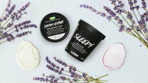 Dream Cream and Sleepy body lotion lie on a pale blue background, surrounded by dried lavender stems and a dollop of each product next to each pot
